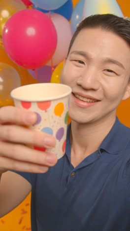 Vertical-Video-Studio-Portrait-Of-Man-Wearing-Party-Hat-Celebrating-Birthday-Doing-Cheers-To-Camera-With-Paper-Cup-With-Balloons